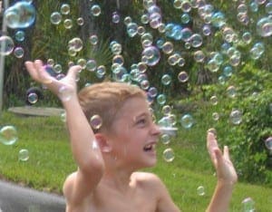 A young child smiles with arms raised amidst a shower of iridescent bubbles.