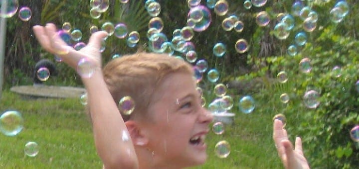 A young child smiles with arms raised amidst a shower of iridescent bubbles.