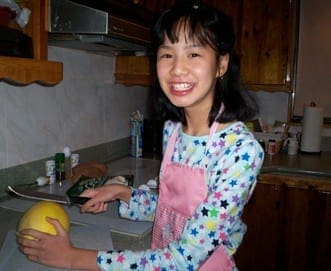 A young girl with dark hair and a pink apron smiles while holding a yellow fruit in a kitchen.