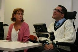 A woman in glasses and a pink cardigan sits with a man in a wheelchair wearing glasses and a sweater vest.