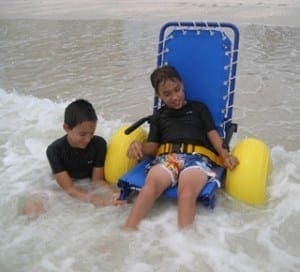 Two boys enjoy the ocean surf with a child in a blue and yellow beach wheelchair.