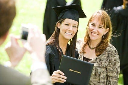 A graduate smiles with her proud mother as a person in the foreground takes a photo.