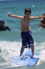 A young boy with arms outstretched rides a surfboard on a wave, with two other people in the background in the ocean.