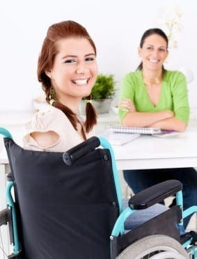 Young woman in a wheelchair smiles at the camera while another woman sits at a desk in the background.