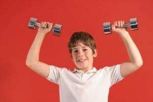 A boy in a white shirt lifts two dumbbells overhead against a red background.
