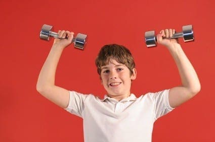 A boy in a white shirt lifts two dumbbells overhead against a red background.