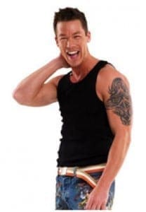 Man with dark hair and a black tank top laughs with his arm behind his head.