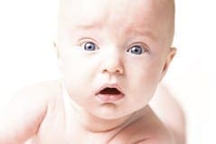 A baby with wide blue eyes and an open mouth looks at the camera with a surprised expression.