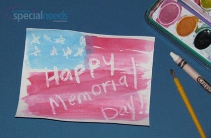 Child's drawing of a flag with "Happy Memorial Day!" written in white crayon, next to art supplies.