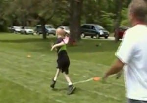 Child in black and green shirt throws a ball on a grassy field while an adult watches.