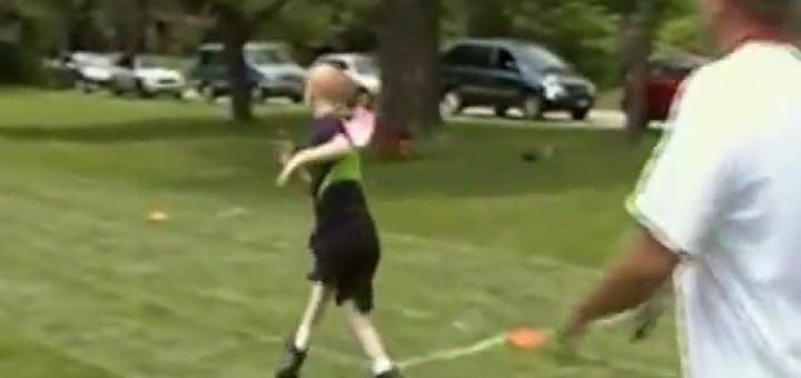 Child in black and green shirt throws a ball on a grassy field while an adult watches.