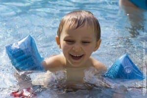 A smiling boy wearing blue arm floaties splashes in a swimming pool.