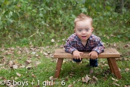 A young boy with Down syndrome smiles from atop a small wooden table outdoors.