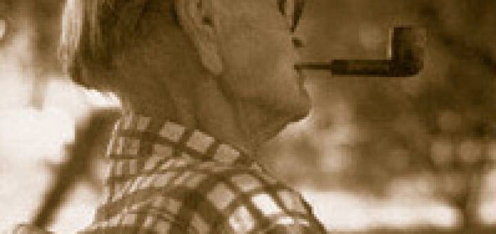Frail elderly man in a plaid shirt and glasses sits alone while smoking a pipe.