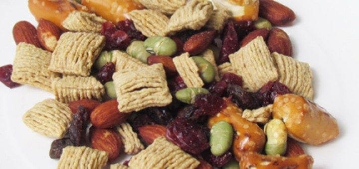 A pile of trail mix containing cereal squares, almonds, pretzels, raisins, cranberries, and edamame.