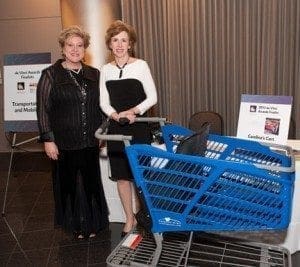 Two women stand next to a blue shopping cart with signs in the background.