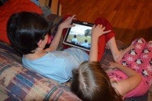 Two children watch a tablet computer on a couch.
