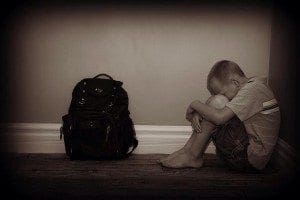 A young boy sits huddled next to a backpack, his face buried in his knees.
