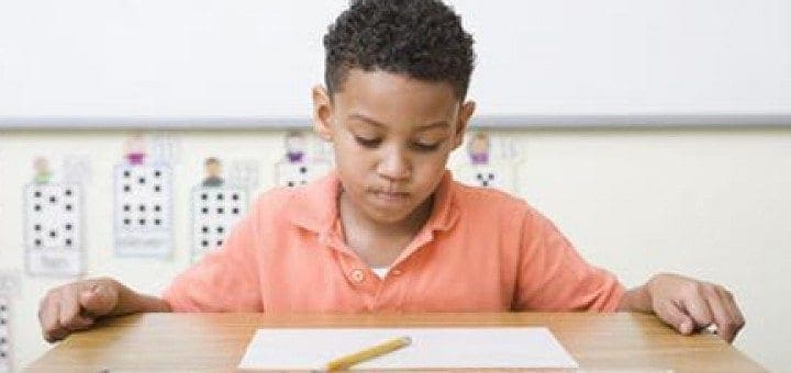 A young boy with dark curly hair sits at a desk with a piece of paper and a pencil.