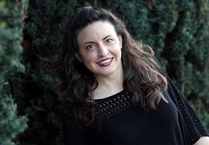 Nina G, a comedian and activist, smiles outdoors with trees in the background.
