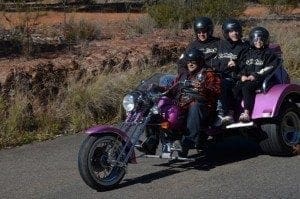 Four people wearing helmets ride a purple three-wheeled motorcycle on a road.