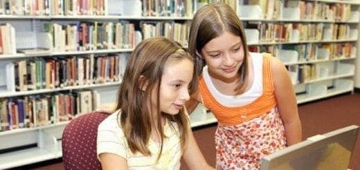 Two girls look at a laptop in front of a bookshelf.