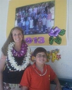 Teacher Megan Rex wearing a floral lei stands smiling beside her student Noah Rubino in front of a 2013 class banner.