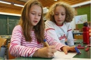 Two young girls work together on a paper at a table in a classroom.