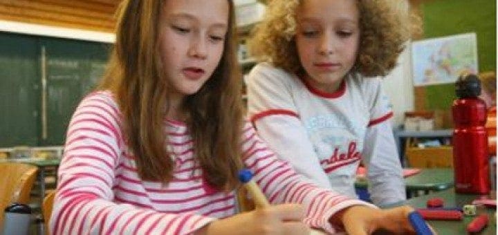 Two young girls work together on a paper at a table in a classroom.