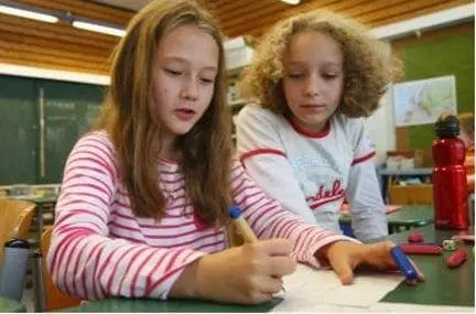 Two young girls work together on a paper at a table in a classroom.