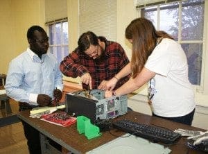 Three people work on disassembling a computer at a lab table.