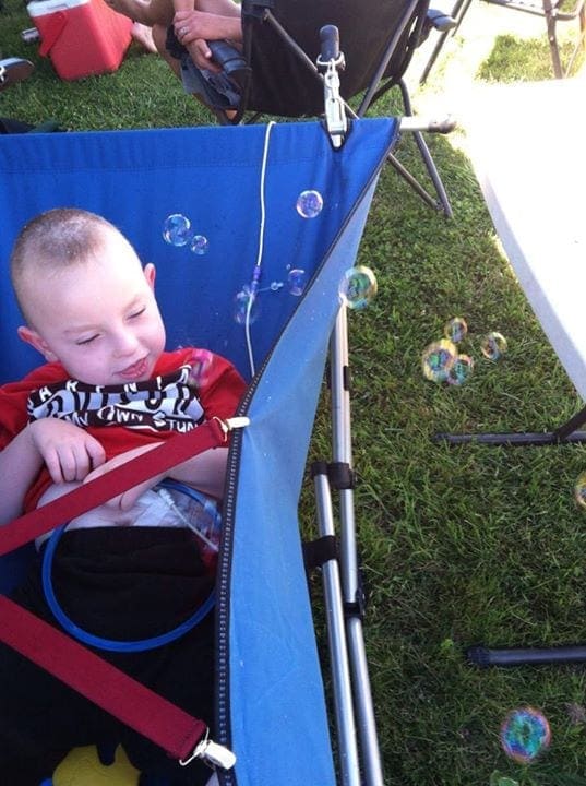 Child in a blue travel bed with straps, watching bubbles float by outdoors.