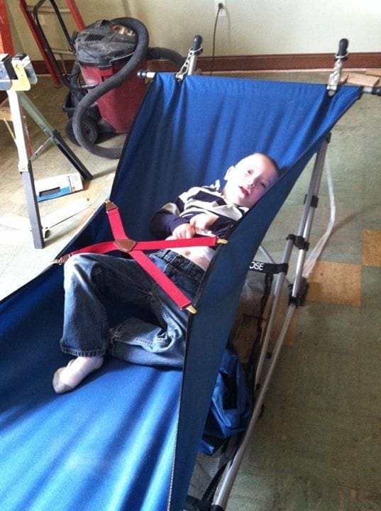 Child lies in a blue travel bed made from a camping chair with added foam and restraints.