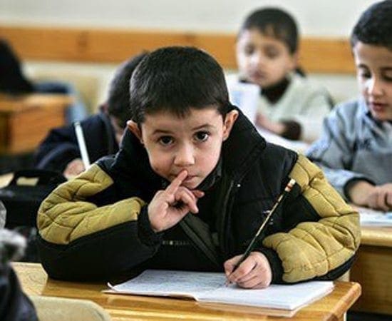 Boy with finger to his lips, sitting at a desk with a pencil and notebook.