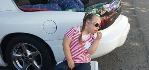 Young girl with braids and sunglasses eats a snack next to a white car.