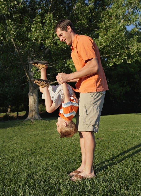 Man holding a child upside down outdoors on a grassy lawn.