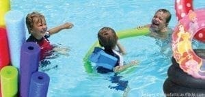 Children play in a swimming pool with pool noodles and inner tubes.