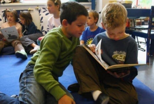 Two young boys read a book together while other children sit in the background.
