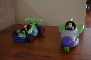 Two Toy Story themed toys, a green race car and a gray and purple airplane, sit on a wooden surface.