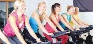 Five women smiling while riding stationary bikes in a fitness class.
