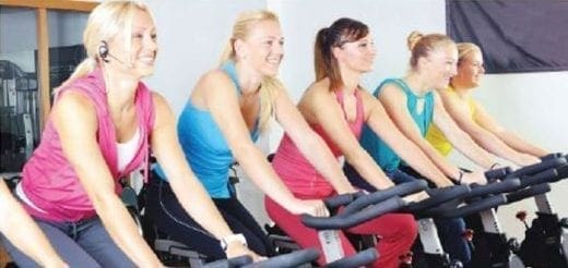 Five women smiling while riding stationary bikes in a fitness class.