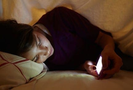 Teenager lying in bed, looking at a glowing smartphone screen in the dark.