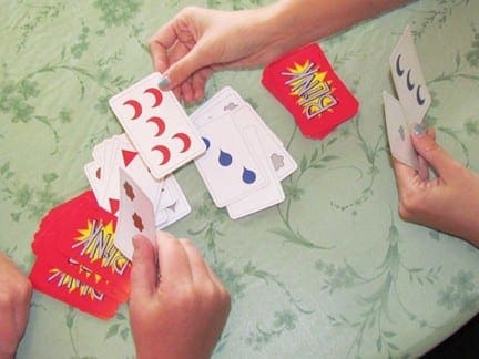 Children playing a card game with red and white cards scattered on a green patterned tablecloth.