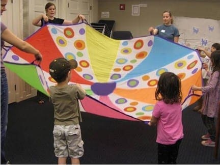 Children and adults hold a colorful parachute with circles during a group activity.