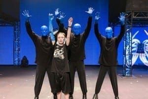 Blue Man Group performers stand with a child on stage with their hands raised.