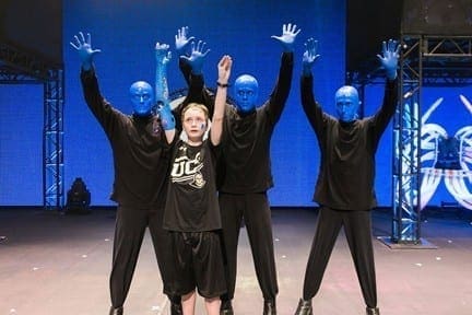 Blue Man Group performers stand with a child on stage with their hands raised.