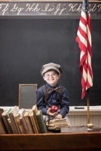 Young boy in vintage clothing sits at a desk with books, an apple, and an American flag in front of a chalkboard.