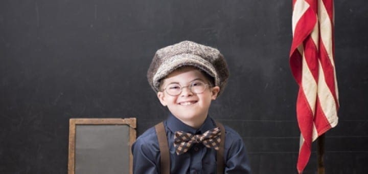 Young boy in vintage clothing sits at a desk with books, an apple, and an American flag in front of a chalkboard.