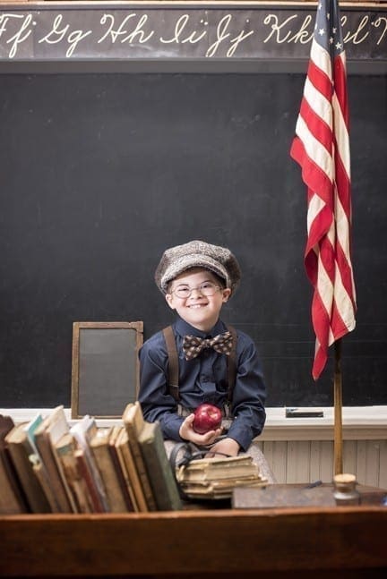 Young boy in vintage clothing sits at a desk with books, an apple, and an American flag in front of a chalkboard.