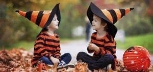 Two children in striped shirts and witch hats sit on the ground surrounded by autumn leaves.